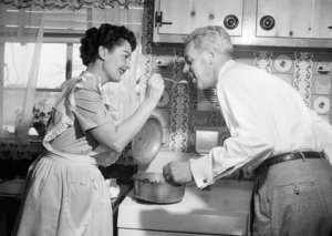 1950s HOUSEWIFE IN KITCHEN HAVING HUSBAND TASTE FOOD ON STOVE
